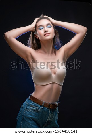 Beautiful mixed race Asian girl with big breasts, wearing a bra and jeans, smiles and touches her hair with her hands. Advertising, commercial design