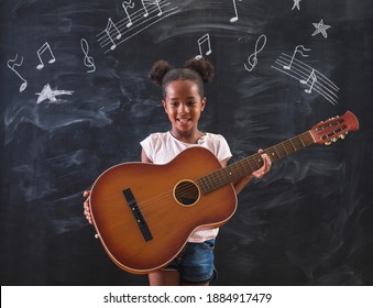 Beautiful Mixed Race Elementary School Girl Standing In Front Of A Chalkboard In Classroom, Playing The Guitar And Singing While In Music Class, With Musical Note Symbols Drawn On The Blackboard