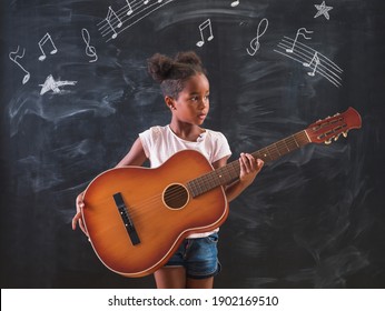 Beautiful Mixed Race Elementary School Girl Standing In Front Of A Chalkboard In Classroom, Playing The Guitar And Singing While In Music Class, With Musical Note Symbols Drawn On The Chalkboard