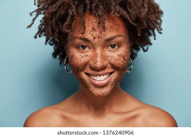 Beautiful mixed race female has skin scrub on face, smiles gently and looks directly at camera, makes cosmetic masks from coffee, has curly hairstyle, bare shoulders, isolated over blue background