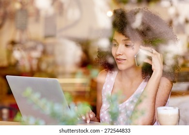 Beautiful Mixed Race Woman Talking On Her Phone While Typing On Her Laptop, Seen Through The Window Of A Coffee Shop       