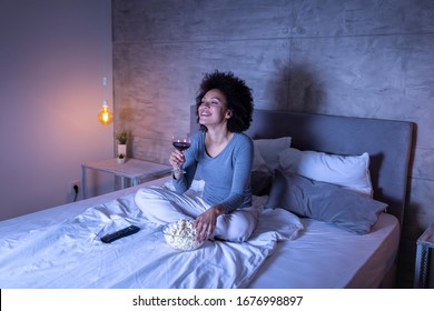 Beautiful Mixed Race Woman Wearing Pajamas Sitting On Bed, Eating Popcorn, Drinking Wine And Watching A Comedy Movie On TV, Relaxing At Home At Night