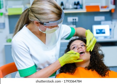 Beautiful Mixed-race Teenage Girl On Dental Checkup By Middle-age Caucasian Woman Wearing Face Mask And Visor As Coronavirus Safety Precaution. Modern Dentist's Office. 