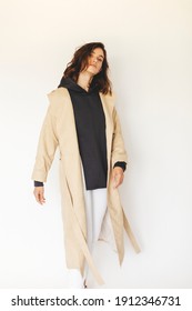 Beautiful Model Turn Around. Sensual Brunette Wear Minimalistic Outfit. Young Woman In Beige Trench Coat, Grey Hoody And White Pants. Winter, Fall Autumn Or Spring Minimal Outfit. Woman Look Elegant.