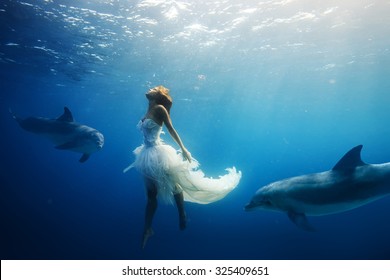 Beautiful model in white dress underwater. A girl diving with dolphins without scuba gear. Fantasy mermaid in deep ocean. Water surface with sunbeams.