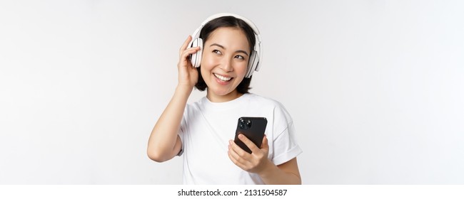 Beautiful modern asian girl, listening music in headphones, holding mobile phone, using smartphone app, standing over white background - Powered by Shutterstock