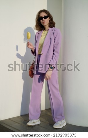 Beautiful modern woman with an ice cream. Fashionable. Violet suit. Yellow top. Brown hair. Black sunglasses. Slim and tall. Daily life. Work and leisure. City background. Bright colors.