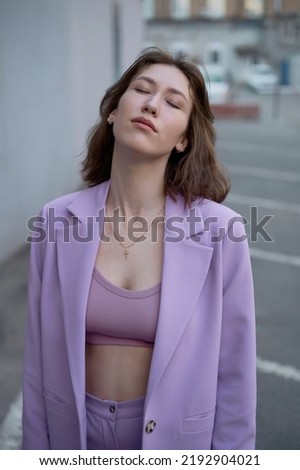 Beautiful modern woman in parking area. Fashionable. Violet suit. Yellow top. Brown hair. European type. Slim and tall. Daily life. Work and leisure. City background. Soft light.