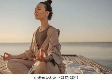 Beautiful modern young woman meditates with closed eyes, relaxes her body and clears her mind. Brown-haired woman with bun on her head is dressed light brown jacket and pants. Rest time concept.