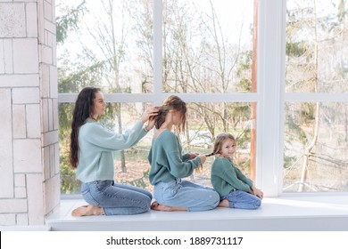 beautiful mom braids hair to older daughter, daughter braids hair to sister. They are sitting on the windowsill. Dressed in the same sweater - Powered by Shutterstock