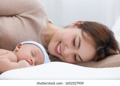 Beautiful Mom Lying At The Back Of The Baby While The Infant Is Sleeping On The White Bed. Asian Mother Looking At The Baby Which Is Her Child With Love And Care.