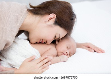 Beautiful Mom Supports And Tenderly Cuddles The Newborn Baby Gently While The Infant Is Sleeping On The White Bed. Asian Mother Kissing And Touching On The Back Of Babies With Love And Care.