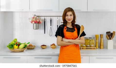 Beautiful Mom Wears Orange Apron Smiling And Confidently Standing With Crossed Arms In Kitchen And Holding Wooden Ladle As Ready To Cook