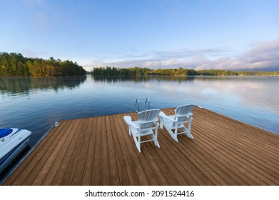Beautiful morning landscape panorama, under a blue sky, of a lake in cottage country in Ontario, Canada. Two white Adirondack chairs facing the calm water are sitting on a new wooden dock. - Powered by Shutterstock