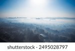 A beautiful morning view of a cityscape covered in fog and mist, with silhouettes of buildings and trees emerging through the haze. The serene and calm atmosphere is enhanced by the soft sunlight filt