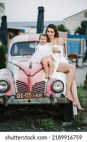 A Beautiful Mother With A Daughter In A White Dress Posing Near Red Retro Car. Mom With Child Enjoying Day. Summer Family Photo. Spring Fashion Model Concept. Vintage And Retro Style. Luxury Travel.