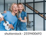 Beautiful mother and her cute little daughter with microphone singing together and playing ukulele. Young caucasian woman and preteen girl enjoying and playing music in the living room at home .