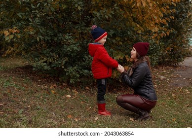 Beautiful Mother Holds The Hands Of Her Son In A Red Jacket And Red Boots In The Park In Autumn. Family Trust Care Concept. A Walk With A Child In Autumn, Boundaries And Rules For Raising Children.