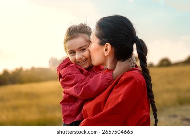 A beautiful mother holds her young daughter and gives her an affectionate kiss on the cheek, while the happy child smiles at the camera, all in a warm sunset field.