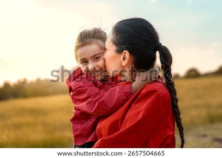 A beautiful mother holds her young daughter and gives her an affectionate kiss on the cheek, while the happy child smiles at the camera, all in a warm sunset field.