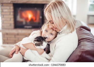 Beautiful Mother, Little Daughter And A Cat Are Sitting On The Sofa Near The Fireplace. Family In The Home Interior. Cozy. Fall.