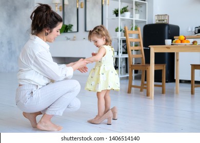 Beautiful Mother And Little Daughter Have Fun In A Bright And Spacious Apartment. The Baby Is Trying On Her Mother's High-heeled Shoes. Mom Is A Young Brunette In White Clothes, A Girl In A Dress.