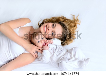 Beautiful mother and newborn baby lying in bed, hugging with love, smiling. Pretty blonde woman and happy infant lay together on white backgound of bed. From high angle shot, above view.