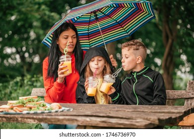 Beautiful mother with two kids are drinking juice at a picnic table in the forest. Happy family having fun together, mother holding umbrella while it is raining. - Powered by Shutterstock