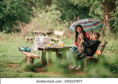 Beautiful Mother With Two Kids Are Drinking Juice At A Picnic Table In The Forest. Happy Family Having Fun Together, Mother Holding Umbrella While It Is Raining. Selective Focus.