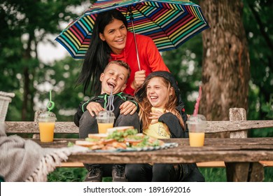 Beautiful mother with two kids are sitting at a picnic table in the forest. Happy family having fun together, mother holding umbrella while it is raining. Selective focus - Powered by Shutterstock