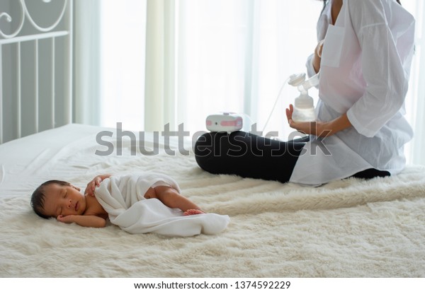 Beautiful mother use automatic breast pump to\
get breast milk and sitting near the sleeping newborn on white bed\
with white curtain  background and day light. Concept of  love and\
warming emotion.