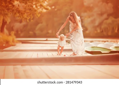 Beautiful Mother Is Walking On The Pier With Her Little Baby Girl Daughter With First Steps At Sunset At Summer Time