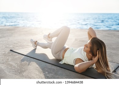 Beautiful motivated young caucasian woman lying quay facing sunrise wearing fitness activewear. Sportswoman doing morning exercise, crunching near sea, improve body shape abs, holding hands behind - Powered by Shutterstock