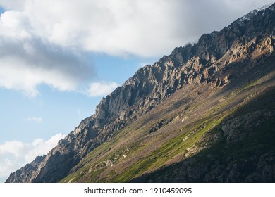 Beautiful mountain landscape with high green mountainside with sharp rocks in sunlight. Atmospheric mountain scenery with steep slope in sunshine. Awesome sunny view to great mountain and low clouds.