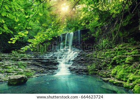 Beautiful mountain rainforest waterfall with fast flowing water and rocks, long exposure. Natural seasonal travel outdoor background with sun shihing
