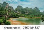 A beautiful mountain range with a lake in the foreground. The lake is surrounded by lush green trees and a few rocks. The sky is clear and blue, and the mountains are covered in trees