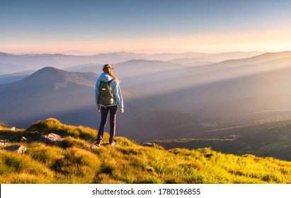 Beautiful mountains in fog and standing young woman with backpack on the peak at sunset in summer. Landscape with sporty girl, green grass, forest, hills , blue sky with sunbeams. Travel and tourism