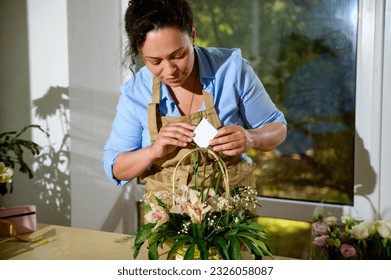 Beautiful multi-ethnic female florist arranging flower composition in wicker basket, putting an empty congratulatory card with free ad space for text. Floral design studio. Flower shop. Small business - Powered by Shutterstock