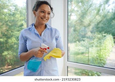 Beautiful Multi-ethnic Woman Housewife, Homemaker, Maid Wearing Blue Casual Shirt, Spraying Detergent On A Yellow Cloth While Washing Panoramic Windows In Veranda. Spring Cleaning And Housekeeping