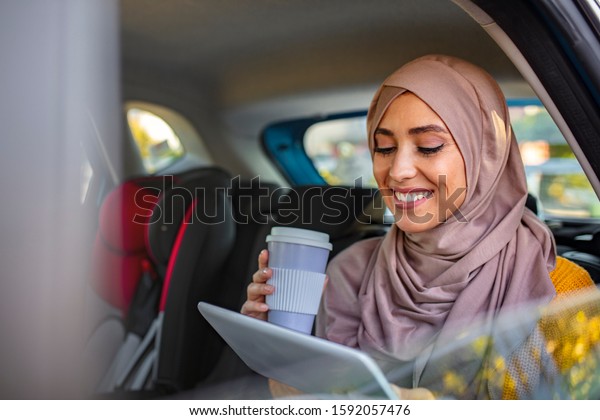 Beautiful muslim arabian woman with hijab sitting on\
backseat in luxury car. She drinking coffee to go and looking at\
tablet computer. Beautiful young Muslim woman in hijab sitting on\
backseat of car