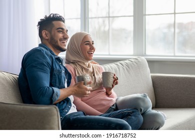Beautiful muslim couple sitting on sofa at home, drinking tea, looking at copy space. Happy young middle-eastern family wife and husband drinking coffee and watching TV, enjoying time together