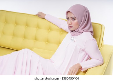 Beautiful Muslim female model wearing modern " baju kurung " dress with hijab sitting on a couch isolated over white background. Eidul fitri fashion and beauty concept. - Shutterstock ID 1380450404