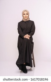Beautiful Muslim female models wearing black long sleeves dress with hijab sitting on a chair, isolated over white background. Stylish Muslim female hijab fashion lifestyle portraiture concept.