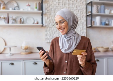 Beautiful muslim girl close up in kitchen, woman in hijab smiling and happy holding smartphone and gold bank credit card, shopping online in online store at home. - Powered by Shutterstock