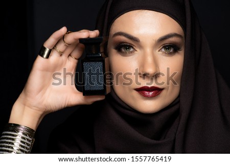 beautiful Muslim woman in hijab with makeup in golden jewelry holding perfume isolated on black