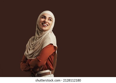Beautiful muslim woman wearing beige hijab, orange blouse and beige pants smiling and looking back on brown background. Copy space. - Powered by Shutterstock