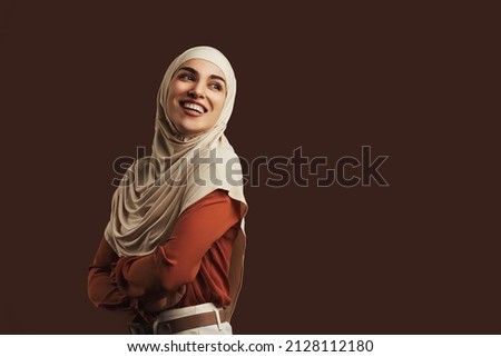 Beautiful muslim woman wearing beige hijab, orange blouse and beige pants smiling and looking back on brown background. Copy space.