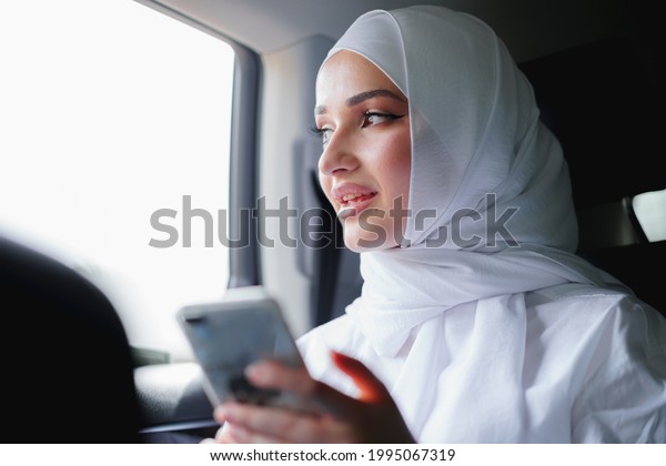 Beautiful muslim woman wearing white\
hijab sitting on the back seat of a car and using\
smartphone
