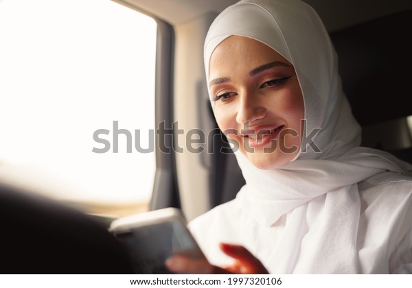 Beautiful muslim woman wearing white\
hijab sitting on the back seat of a car and using\
smartphone