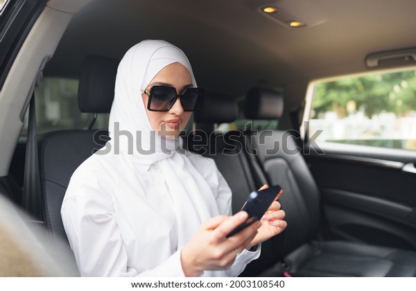 Beautiful muslim woman wearing white
hijab sitting on the back seat of a car and using
smartphone